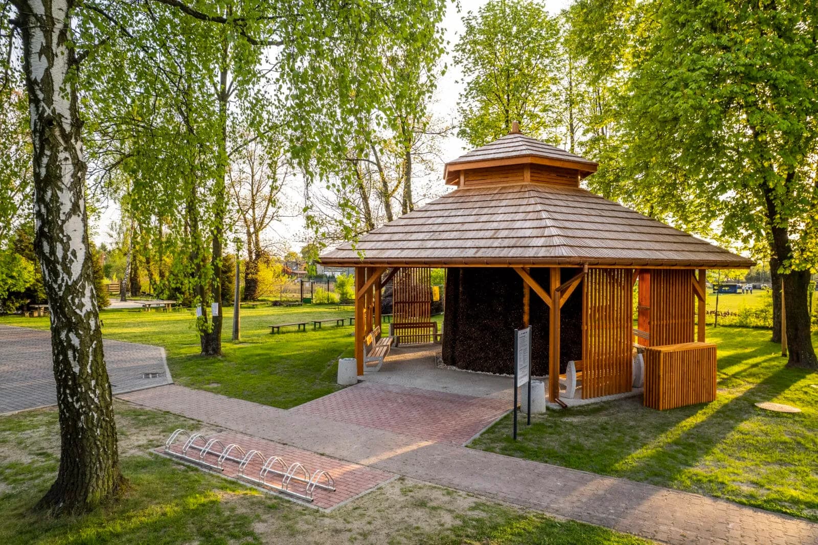A wooden gazebo with a shingled roof stands amidst green grass and trees, with a paved path and bicycle rack in the foreground.